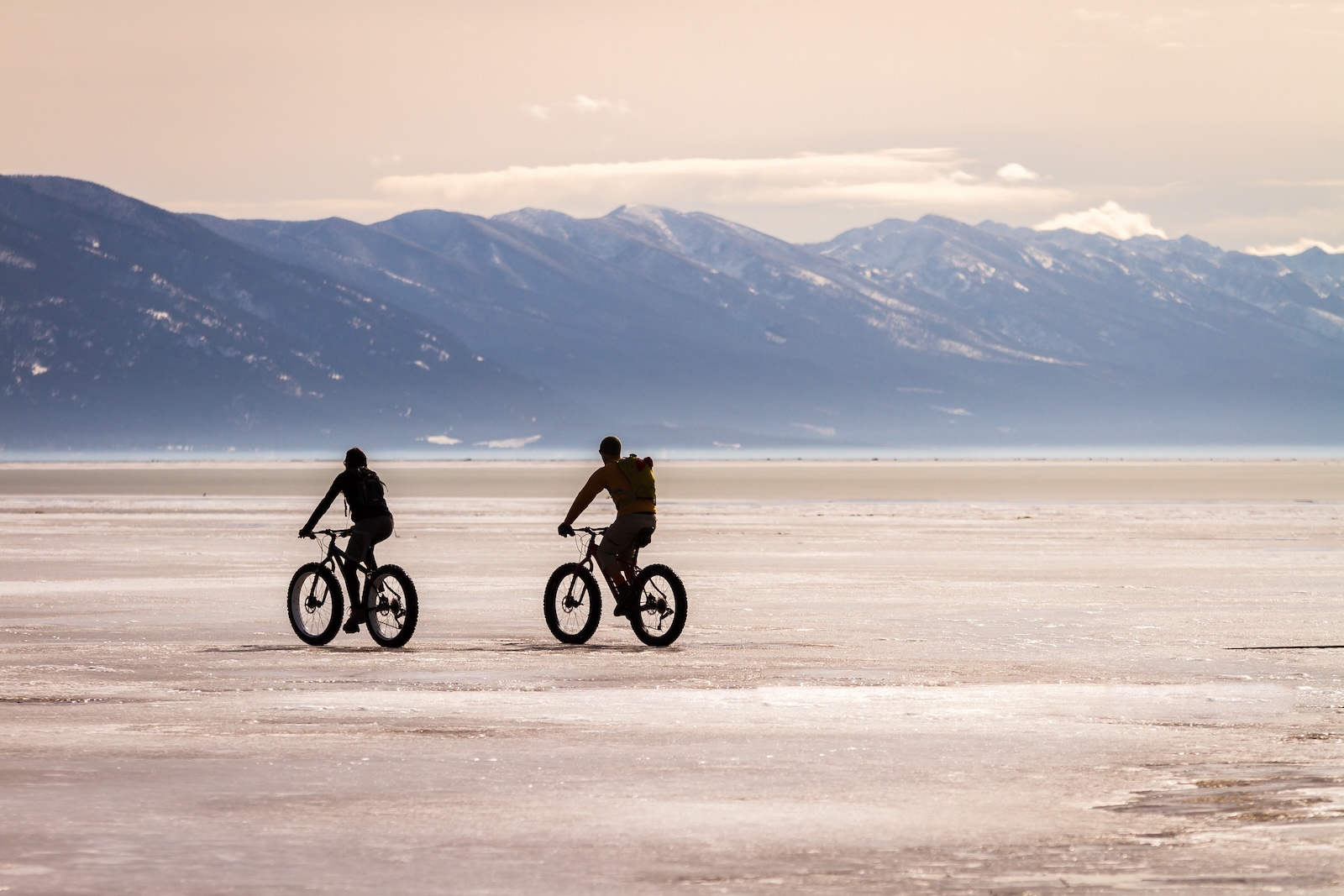 Two bicyclists ride fat tire bikes across a frozen Flathead lake with the Mission Mountain range in the background near Kalispell MT.