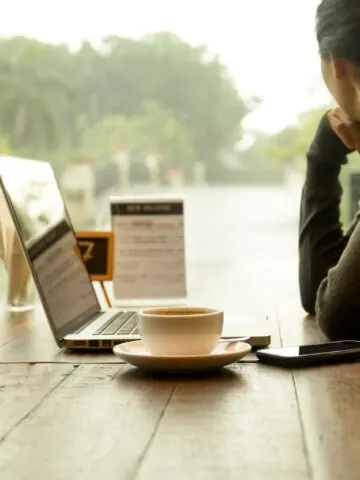 Asian woman with laptop with coffee on the table watching the rain out of window