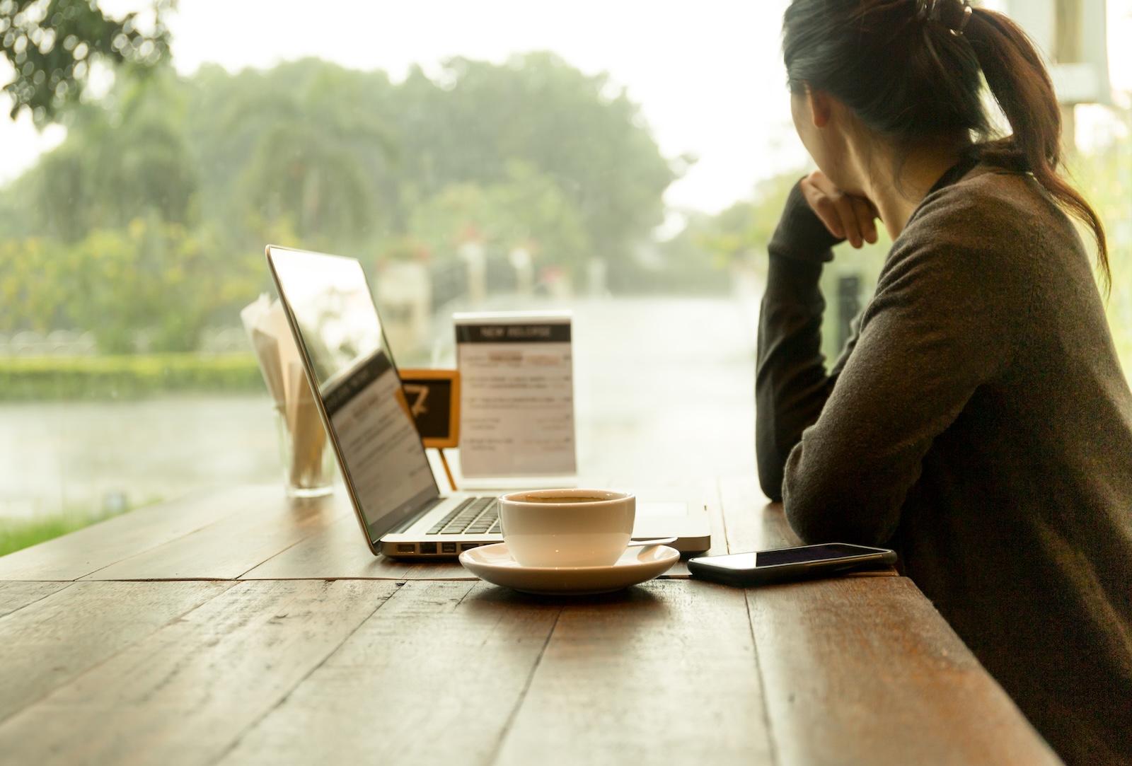 Asian woman with laptop with coffee on the table watching the rain out of window