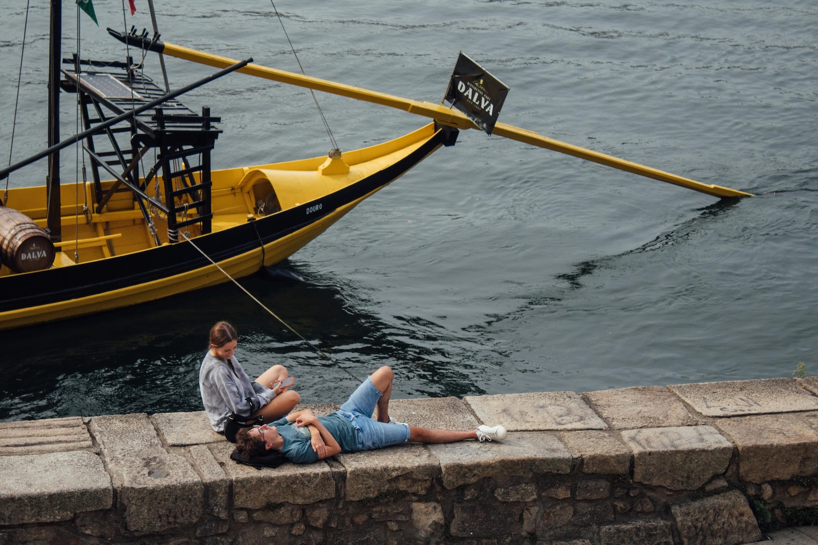 Woman Sitting and Man Lying Down on Wall by River in Porto